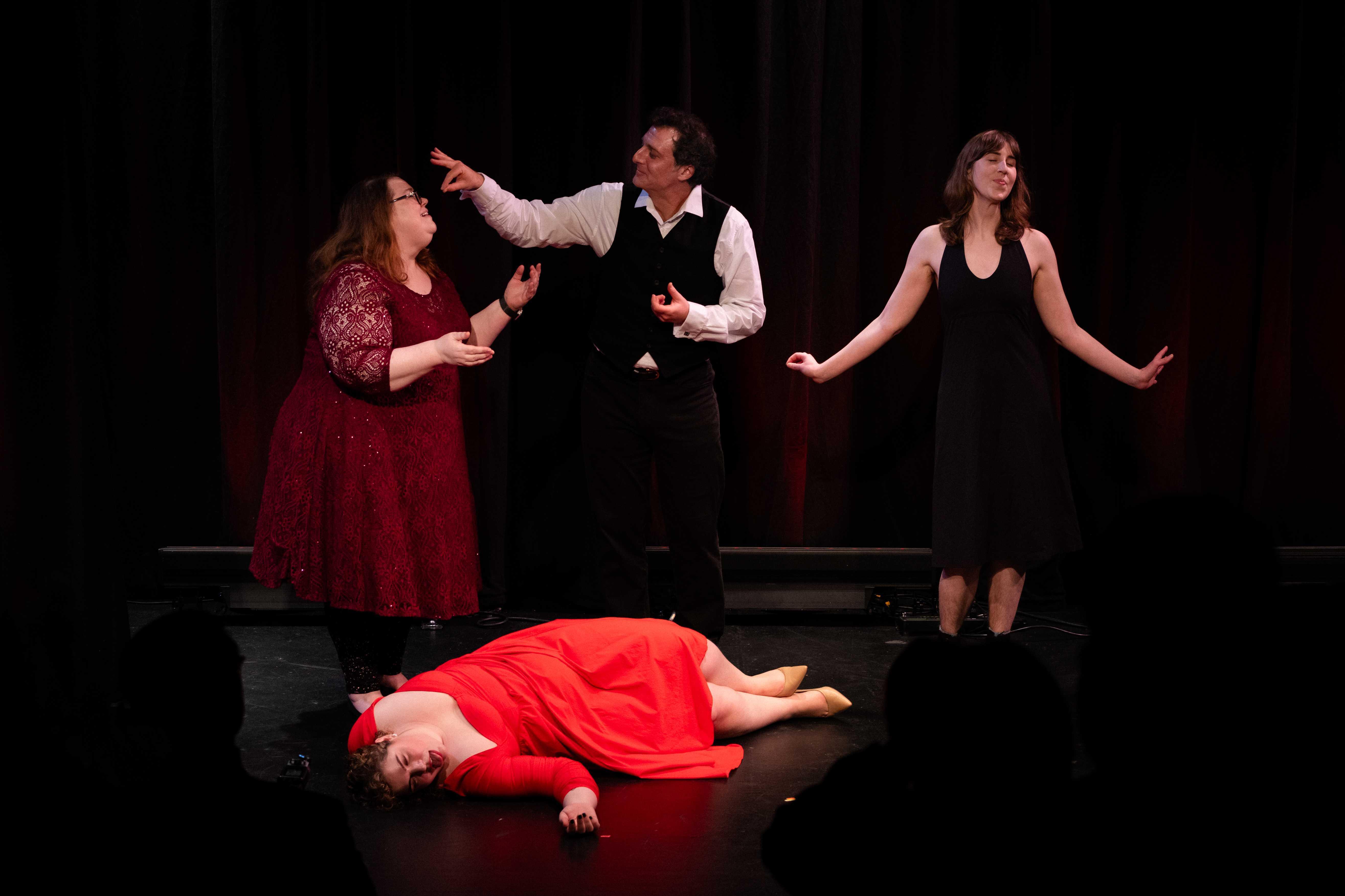 Tableau of actors wearing formal outfits; a man mimes feeding grapes to a woman; a woman in a red dress lies dead on the floor while Kat closes eyes in expression of contentment